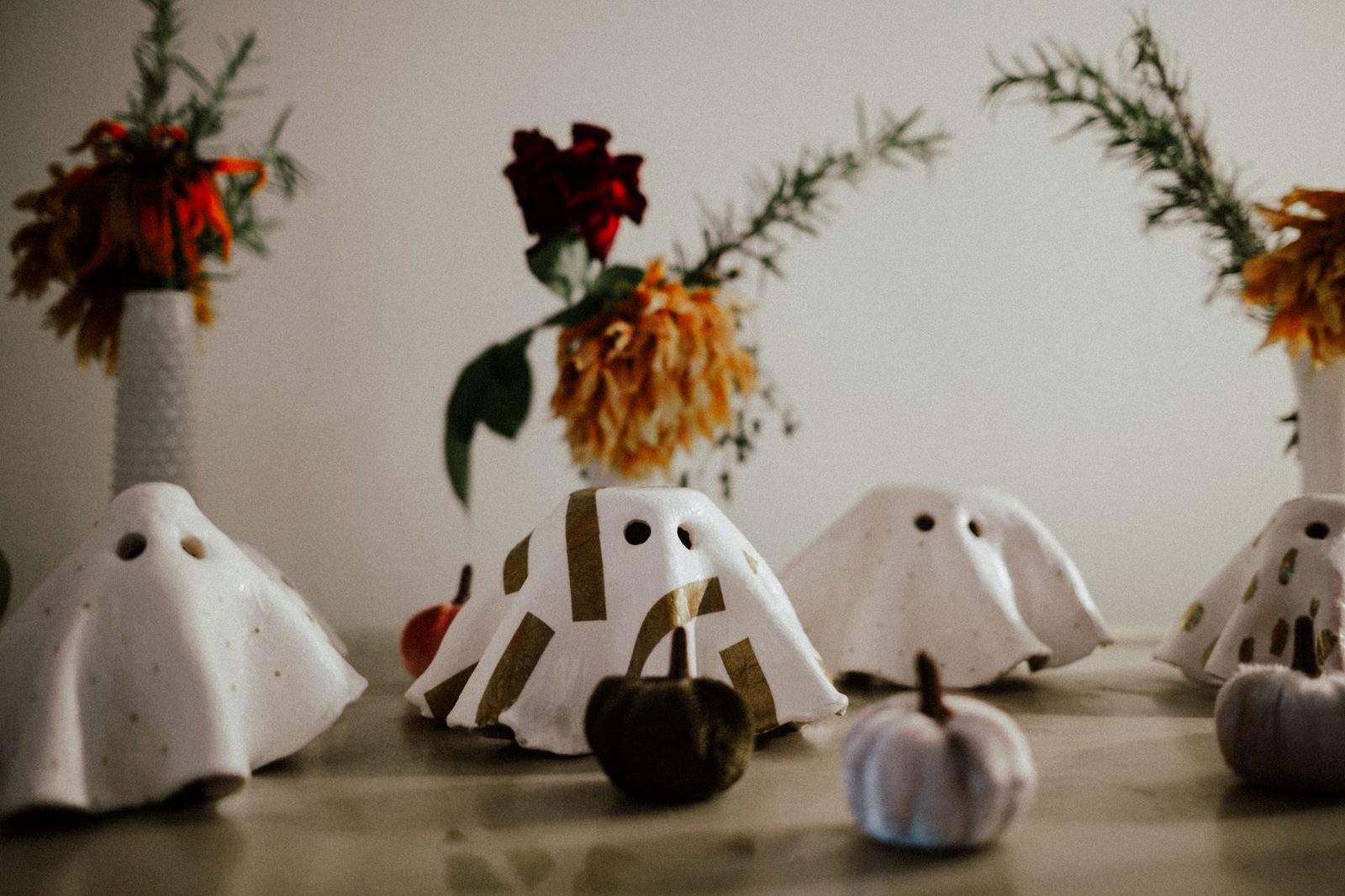 Enfeites de fantasminha branco com olhos vazados e mini abóboras sobre uma mesa, com flores desfocadas ao fundo