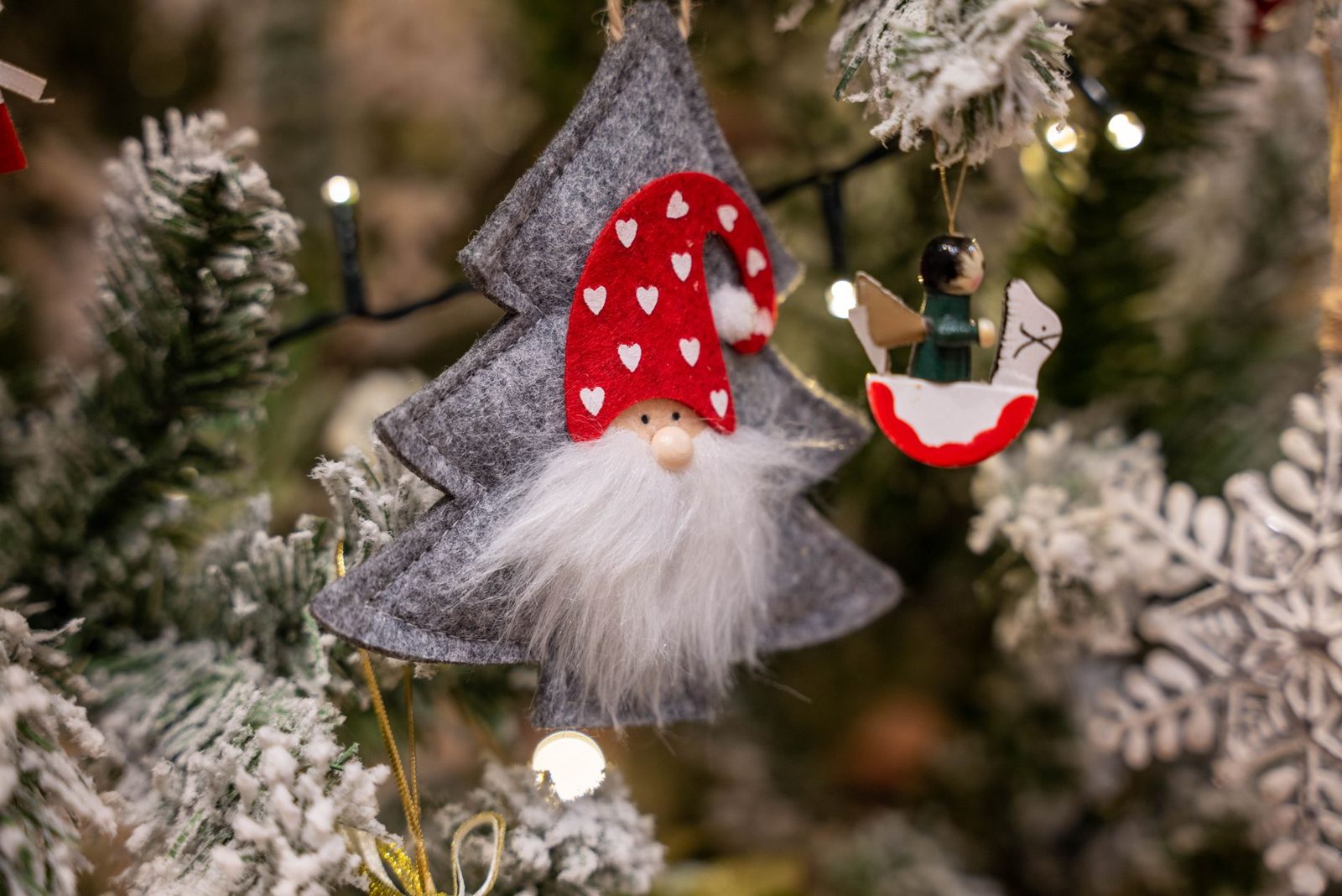 Enfeites de árvore de Natal em feltro — um gnomo de gorro vermelho e um cavalinho de balanço — pendurados em galhos nevados com luzes