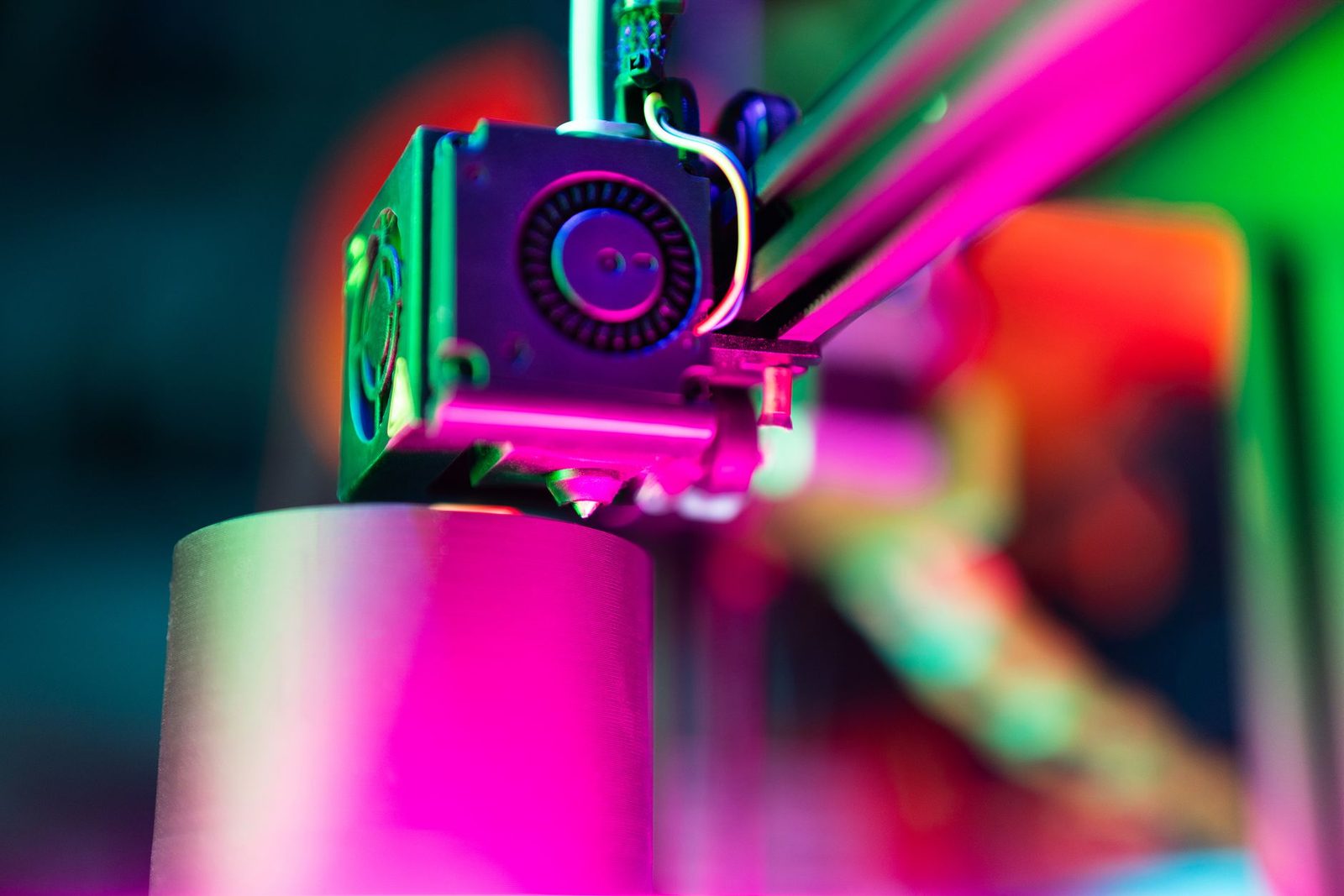 Close do cabeçote de uma impressora 3D FDM imprimindo um cilindro, sob luz neon rosa e verde