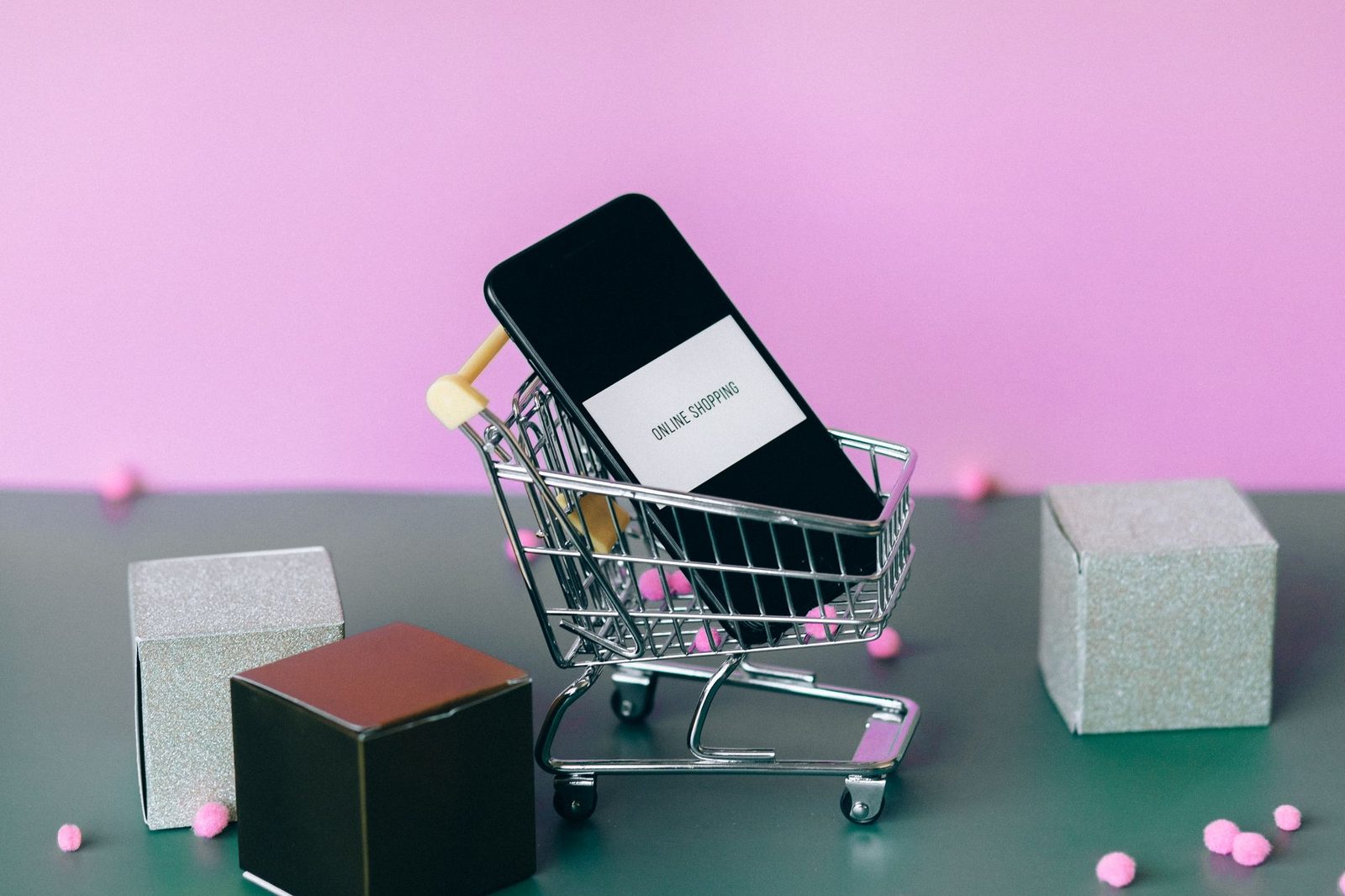 Celular dentro de um carrinho de compras em miniatura, com a tela escrita "online shopping", ao lado de caixinhas de presente sobre fundo rosa