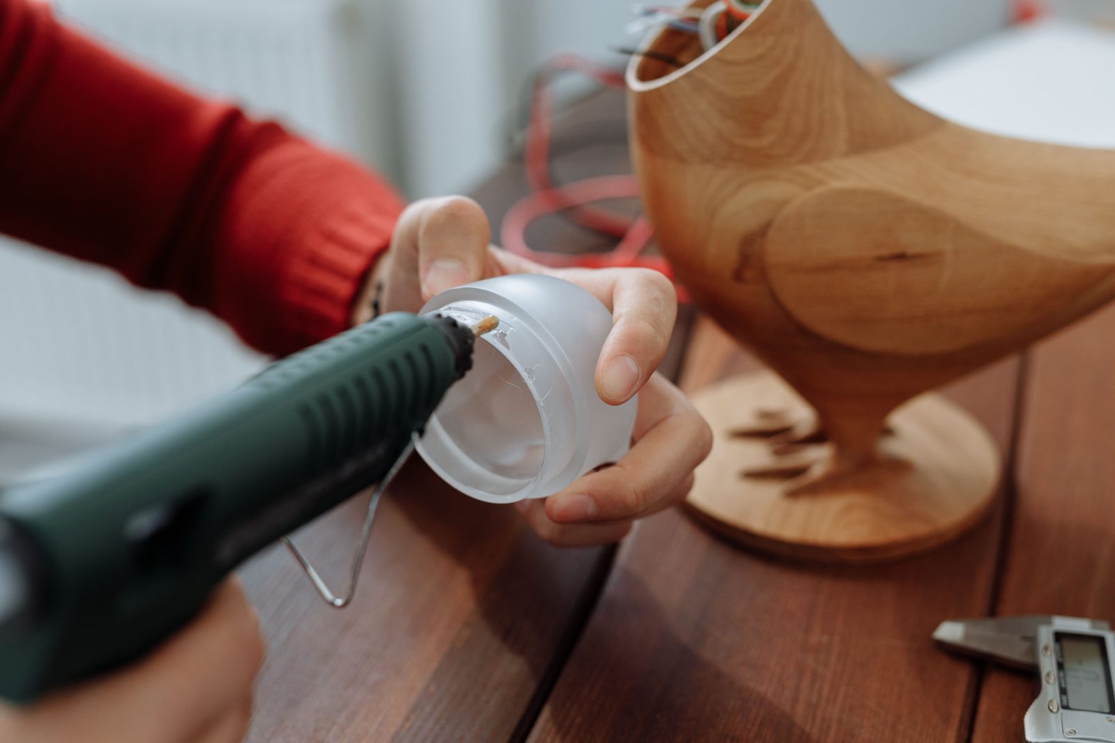 Mão aplicando cola quente com uma pistola verde na borda de uma peça de plástico translúcido, ao lado de um modelo de madeira e um paquímetro digital