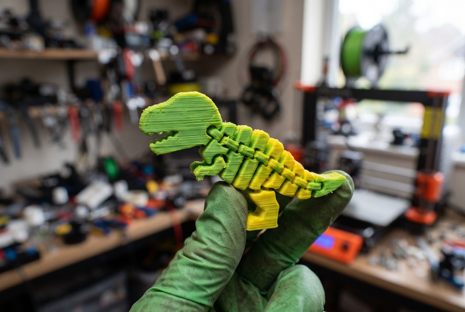 Dinossauro articulado verde e amarelo impresso em duas cores segurado por uma mão com luva