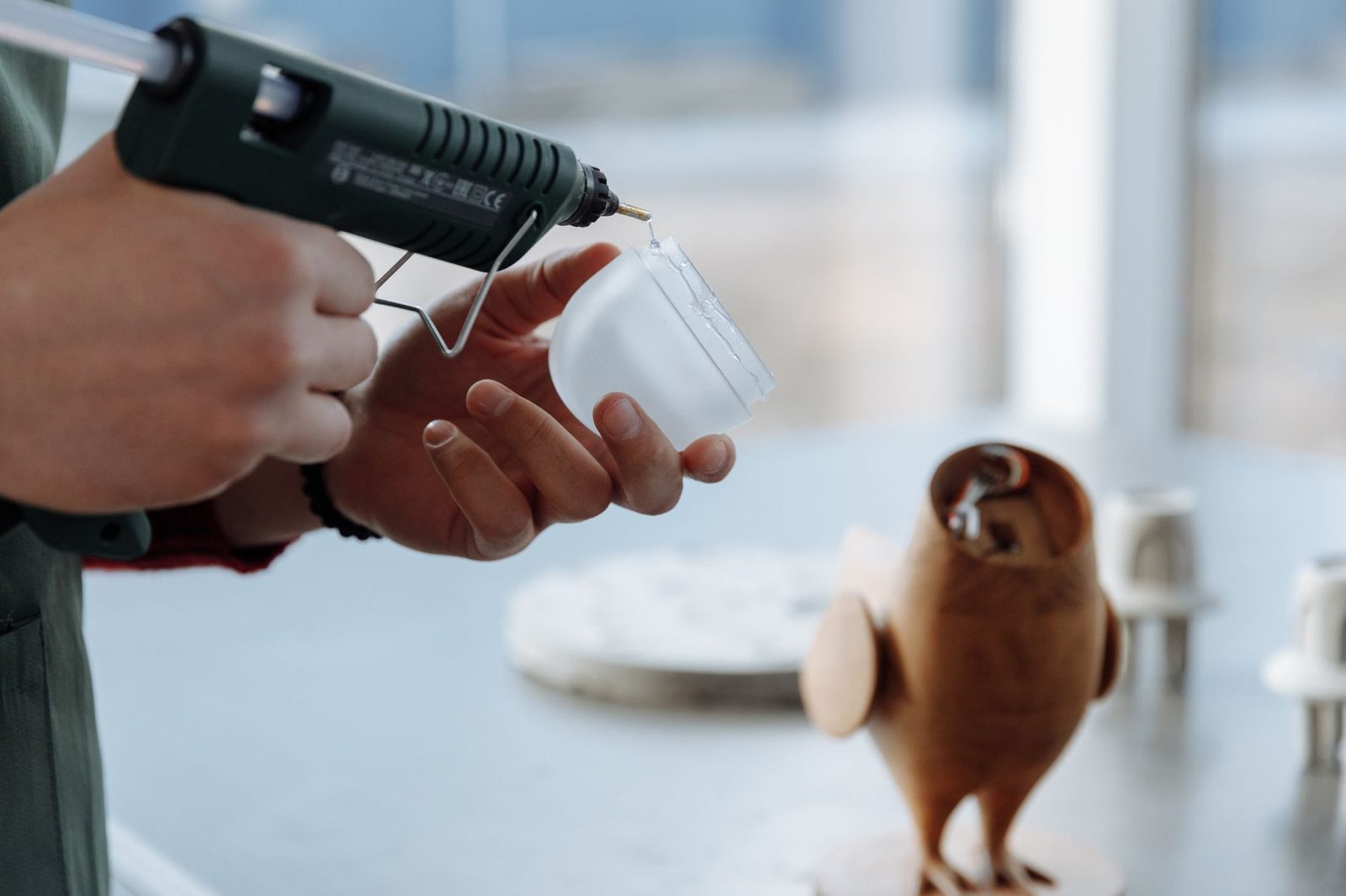 Mãos segurando uma pistola de cola quente sobre uma bancada com bonecos de madeira