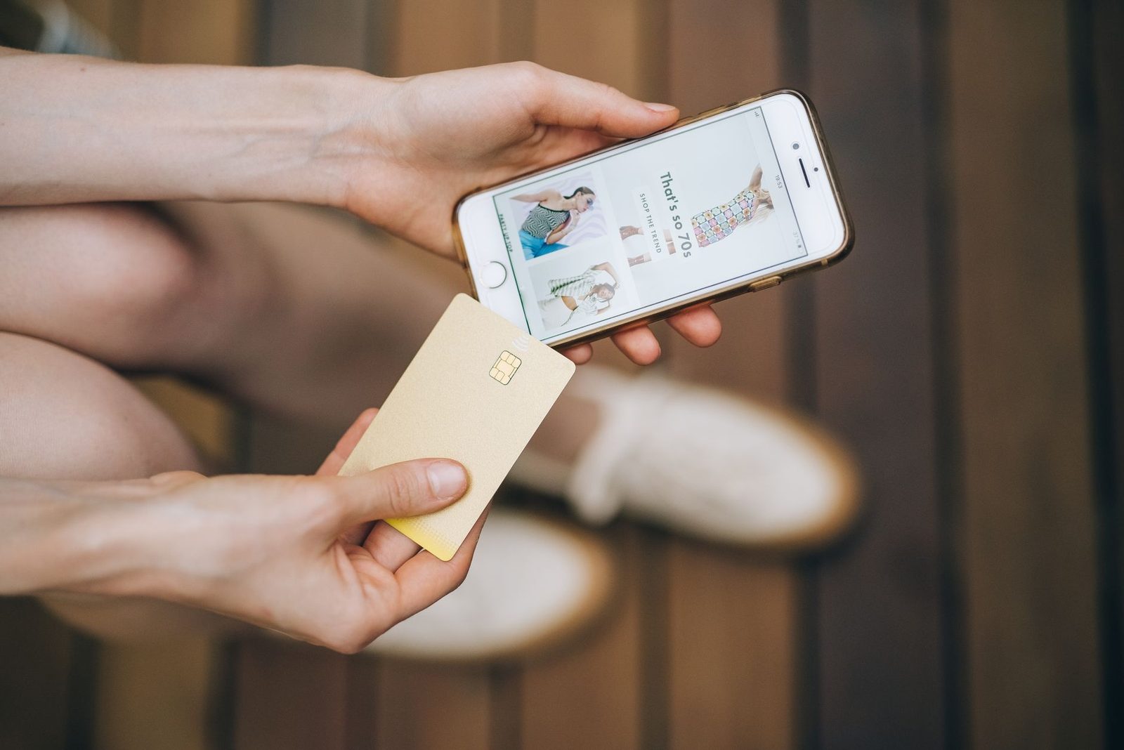 Mãos segurando um celular com uma loja online aberta e um cartão de crédito, vistos de cima sobre piso de madeira