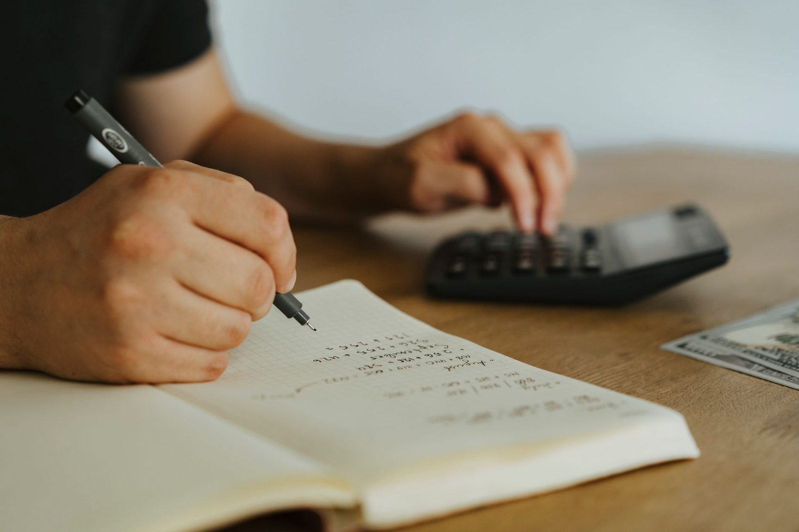 Mão escrevendo contas num caderno aberto, com uma calculadora ao lado sobre a mesa de madeira