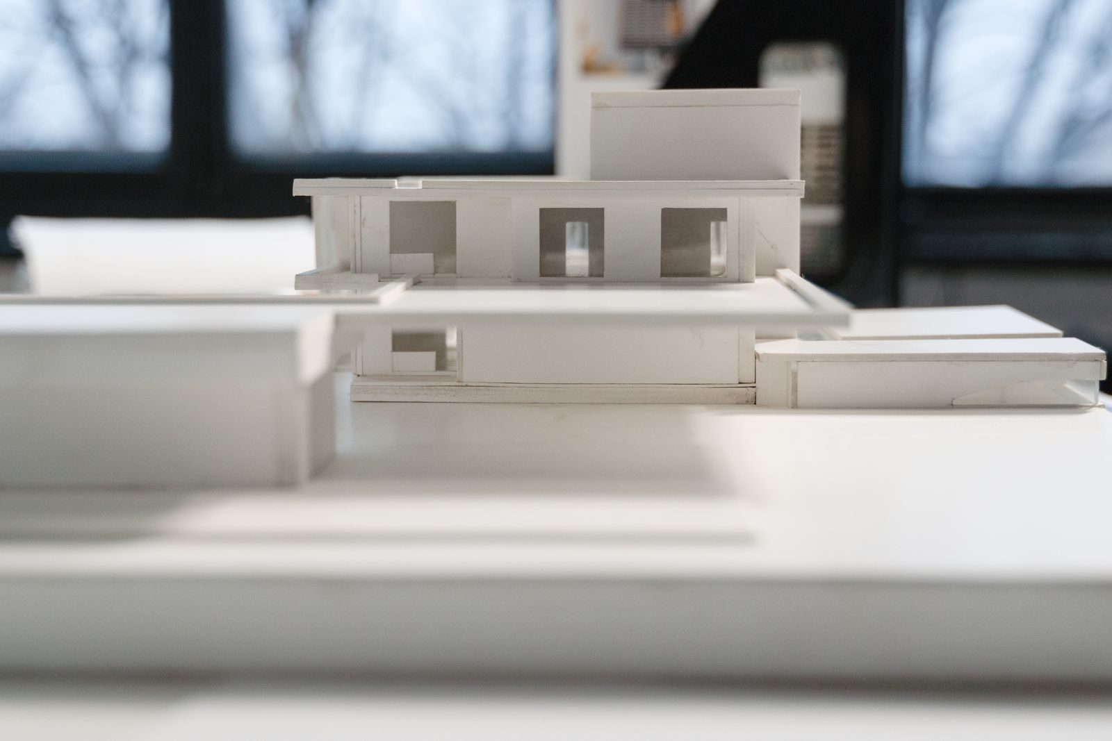 Maquete arquitetônica branca de uma casa de linhas retas sobre uma mesa, com uma janela desfocada ao fundo