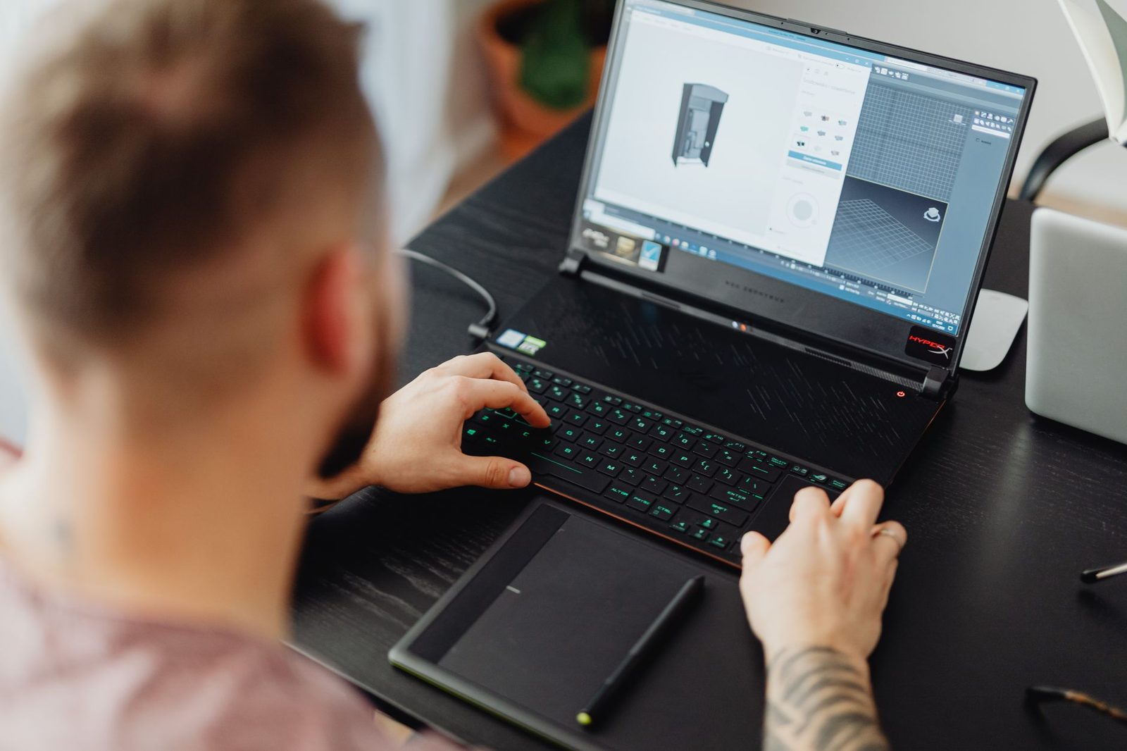 Homem visto por trás do ombro usando um notebook com um modelo 3D aberto num programa de modelagem, com uma mesa digitalizadora ao lado do teclado