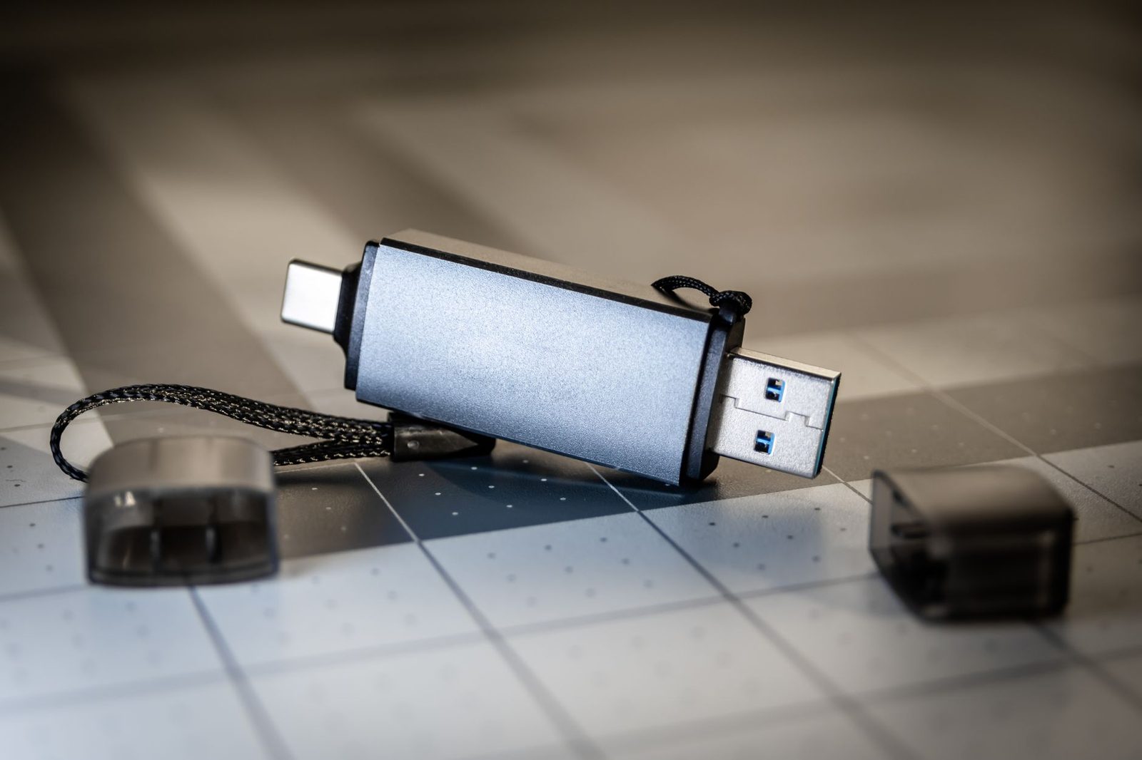 Pendrive prateado com conector USB-A de um lado e USB-C do outro, sobre uma base quadriculada, com as duas tampas soltas ao lado