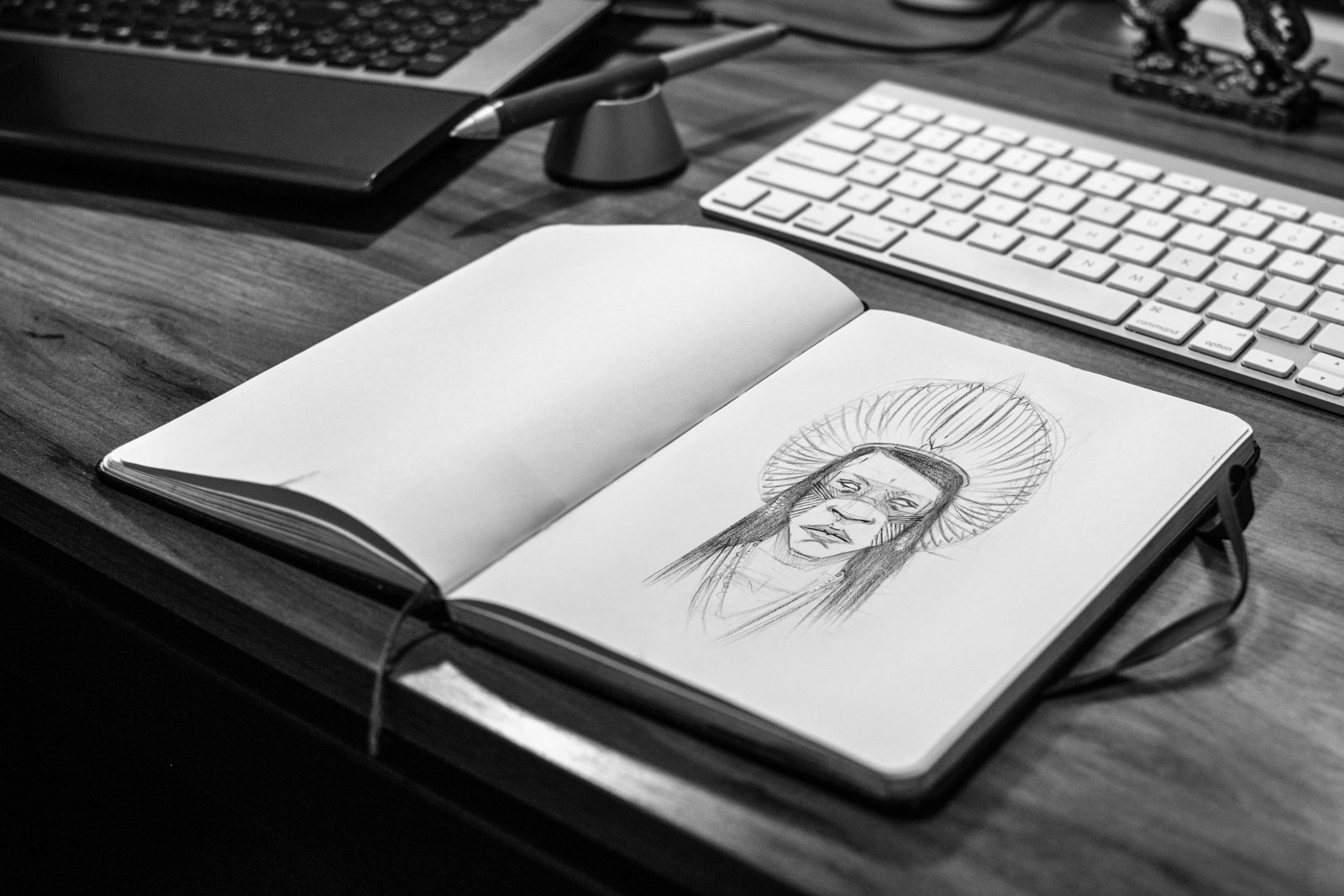 Caderno aberto com um desenho a lápis de um rosto com cocar, sobre uma mesa de madeira ao lado de um teclado branco, em preto e branco